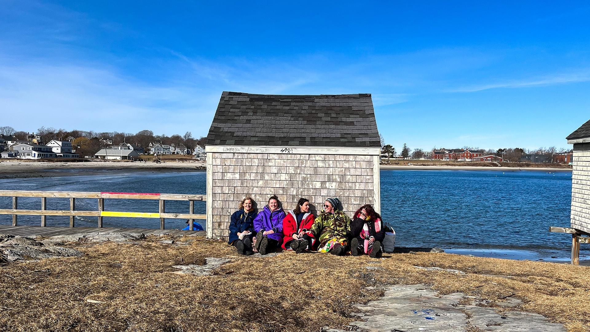 5 women sitting on the ground leaning up against a shed