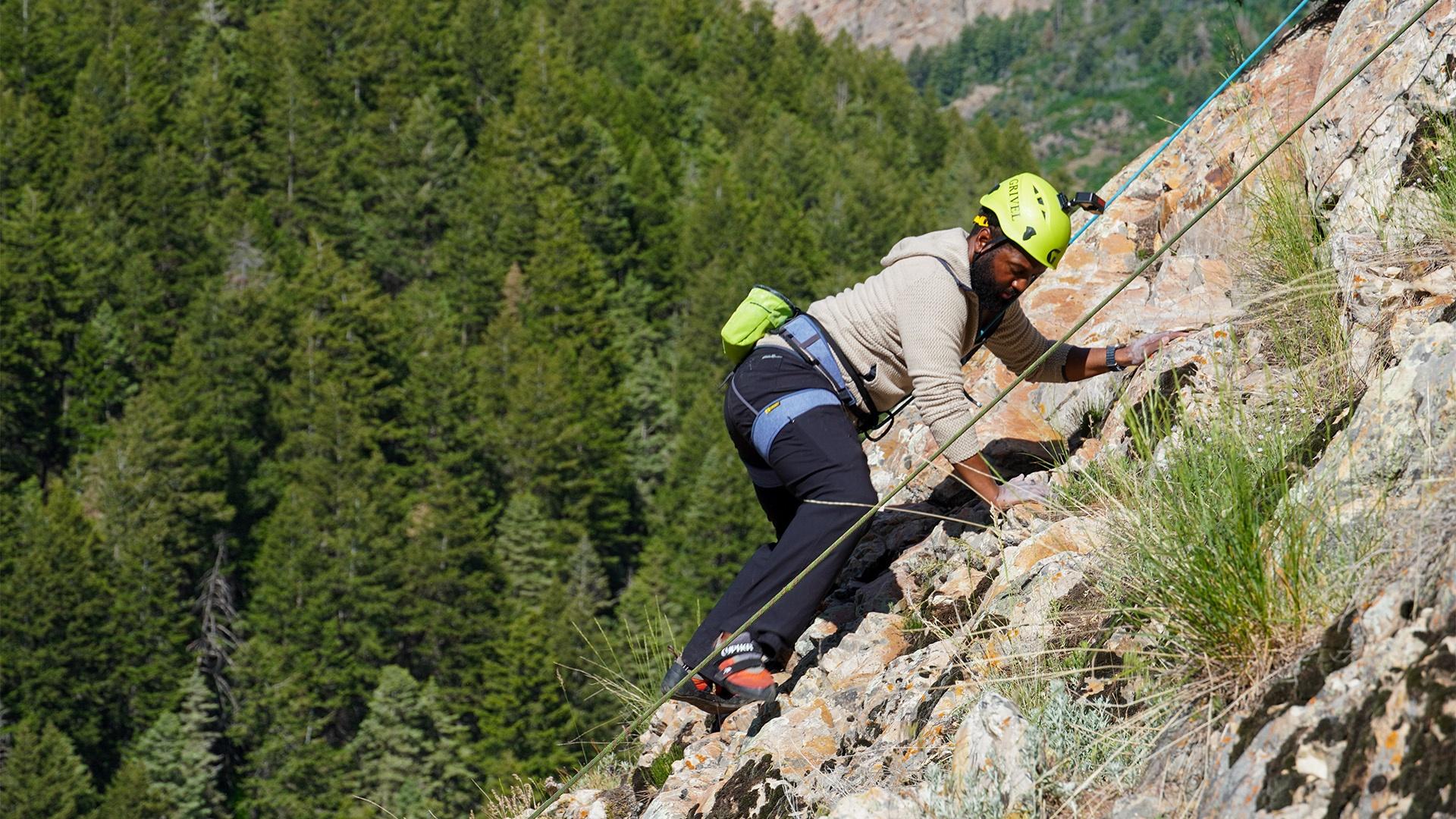 Baratunde climbing up a cliff face with a forest in the background